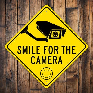 以下が含まれることがあります： 黒いセキュリティカメラのシルエットと「SMILE FOR THE CAMERA」という黒いテキストが書かれた黄色のひし形型の標識。標識の下には黄色のスマイリーマークがあります。