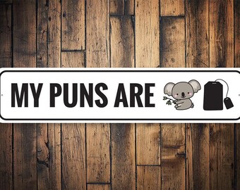 May include: White metal sign with black text that reads "MY PUNS ARE" with a cartoon koala holding a tea bag and a tea bag with a string hanging off of it.