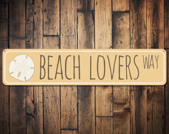 May include: A tan metal sign with a white border and the text "Beach Lovers Way" in brown lettering. A white seashell is on the left side of the sign.