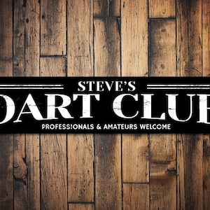 May include: Black and white sign with the text "Steve's Dart Club" and the words "Professionals & Amateurs Welcome". The sign is mounted on a wooden wall.