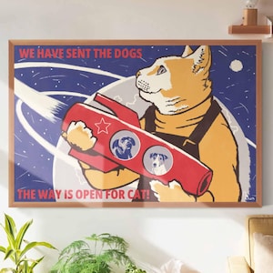 以下が含まれることがあります： 犬の肖像画が描かれた赤いロケットを持つ漫画の猫が描かれた額入りのアートプリント。「WE HAVE SENT THE DOGS」と「THE WAY IS OPEN FOR CAT！」のテキストが青と白の宇宙の背景に表示されています。