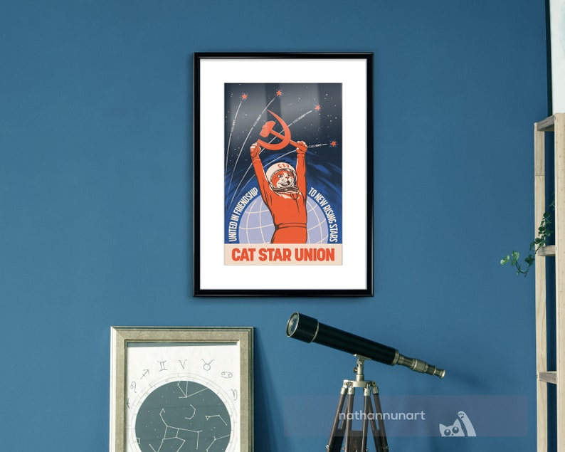 May include: Framed poster with a retro design, depicting a cat in a spacesuit holding a hammer and sickle. The poster's text reads "CAT STAR UNION", with additional text: "UNITED IN FRIENDSHIP" and "TO NEW RISING STARS". The colour scheme is predominantly blue and orange.