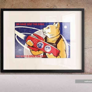 May include: Framed art print with a cartoon cat in a space suit holding a red rocket. The print has the text "WE HAVE SENT THE DOGS" and "THE WAY IS OPEN FOR CATS!" against a blue and white background.