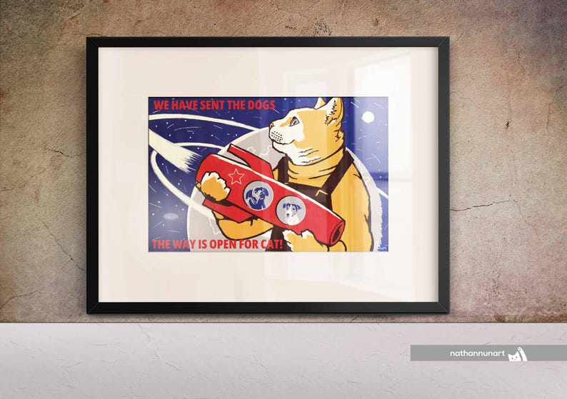 May include: Framed art print with a cartoon cat in a space suit holding a red rocket. The print has the text "WE HAVE SENT THE DOGS" and "THE WAY IS OPEN FOR CATS!" against a blue and white background.