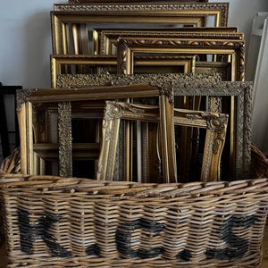 Puede incluir: Una colección de marcos de fotos dorados y ornamentados de varios tamaños y estilos, apilados dentro de una gran cesta de mimbre tejida. Los marcos tienen tallas y adornos detallados. La cesta tiene texto pintado en negro.