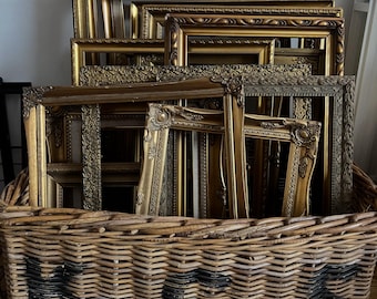 Può includere: Una collezione di cornici dorate e ornate di varie dimensioni e stili, impilate all'interno di un grande cesto di vimini intrecciato. Le cornici presentano intagli e abbellimenti dettagliati. Il cesto ha un testo dipinto di nero.