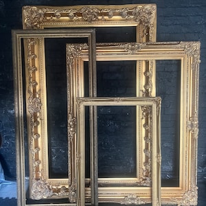 Puede incluir: Cuatro marcos de fotos dorados ornamentados de diferentes tamaños están apilados uno encima del otro contra un fondo de pared de ladrillo negro.