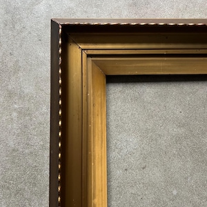 Puede incluir: Primer plano de una esquina de un marco de fotos rectangular. El marco es de madera y tiene un acabado dorado en el interior y un acabado marrón oscuro en el exterior. El marco tiene un borde decorativo.