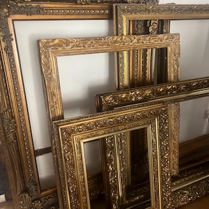 May include: Several antique gold picture frames with elaborate, carved detailing. The frames are of varying sizes and shapes, some with white backing visible. The frames are arranged on a wooden floor, showcasing their intricate designs and golden hue.