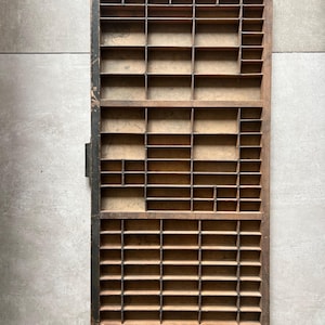 May include: A rectangular wooden printer's type case featuring a grid of numerous small, open compartments of varying sizes. The frame is constructed from dark brown wood with visible signs of wear, including darkened edges and a weathered finish.