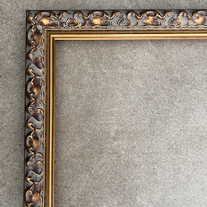 Può includere: Cornice rettangolare ornata con finitura oro e marrone scuro. La cornice presenta un motivo decorativo a volute lungo il bordo esterno e un bordo interno dorato. La cornice è vuota, pronta per una foto o un'opera d'arte.