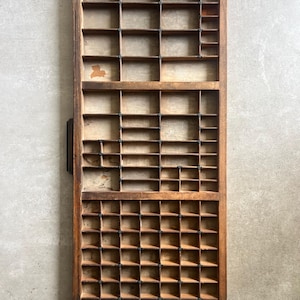 May include: A rectangular wooden printer's drawer featuring a grid of numerous small, open compartments of varying sizes. The wood has a natural, weathered brown finish with visible grain and signs of wear, including small patches of discoloration.