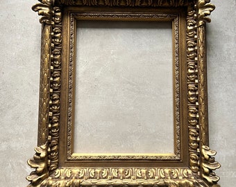 Könnte beinhalten: Ein großer, verzierter Bilderrahmen in Gold mit einem detaillierten, geschnitzten Design. Der Rahmen hat eine rechteckige Form mit einem leicht erhöhten Rand um die Öffnung.