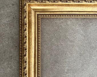 Puede incluir: Un marco de imagen dorado con detalles ornamentados y un borde de cuentas. El marco está contra un fondo gris.