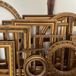 Puede incluir: Una colección de marcos dorados antiguos y ornamentados de varias formas y tamaños. Los marcos incluyen diseños rectangulares y circulares, con tallas detalladas y bordes decorativos. Los marcos están colocados contra una pared blanca y un suelo de madera.