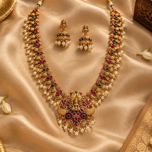 May include: A gold-toned necklace and matching jhumka earring set featuring intricate metalwork, embedded with small red and green stones, and accented with dangling white pearls. The central pendant of the necklace displays a detailed deity figure.