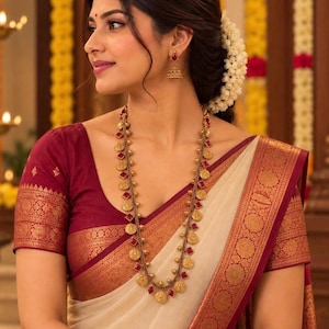 May include: A long gold necklace featuring circular coin pendants and square red stones, paired with matching gold drop earrings. The jewelry is worn with a cream-colored garment featuring a wide, ornate maroon and gold metallic border.