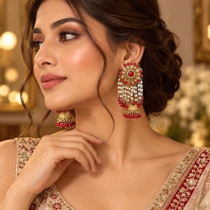 May include: Gold-toned jhumka earrings featuring a circular stud with a central red stone surrounded by clear crystals. The design includes multiple strands of small white pearls and red beads hanging from the stud, ending in a bell-shaped base.