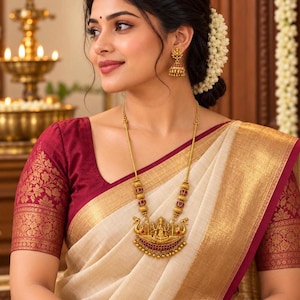 May include: A gold necklace featuring a central pendant with a deity figure flanked by two peacocks, accented with red stones and small gold beads. Matching gold jhumka earrings with red stone details are worn with a cream and maroon saree.