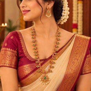 May include: A gold necklace and matching drop earrings featuring green stones, red accents, and small white pearls. The jewelry is worn with a cream-colored garment featuring a wide, deep red border with intricate gold metallic patterns.