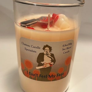 Op de afbeelding: Een glazen kaars pot met een witte sojawas kaars. De kaars heeft een rode en witte afbeelding van een kettingzaag en de tekst "I Can't Feel My Face". Het etiket vermeldt ook "Chateau Candle Specialists" en "6.5oz/185g Soy Blend Candle".