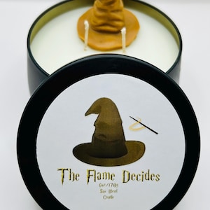 May include: A black candle tin with a white candle inside, topped with a brown Sorting Hat wax figure. The lid features a Sorting Hat illustration and the text "The Flame Decides". The candle is 6oz/170g Soy Blend.