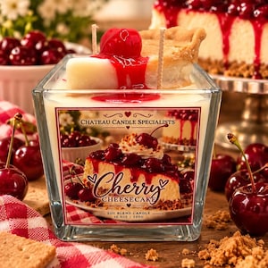 May include: A square glass candle with a soy blend wax top shaped like a slice of cherry cheesecake, featuring a red cherry topping and two wicks. The label reads Chateau Candle Specialists Cherry Cheesecake Soy Blend Candle 10.5oz / 300g.
