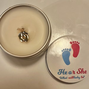 Puede incluir: Una vela blanca en una lata de plata con un dije de pies de bebé de oro. La tapa de la lata tiene un diseño de huellas de pies azul y rosa con el texto "He or She What will baby be?"