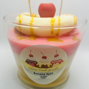 Könnte beinhalten: Eine Glaskerze mit einem geschichteten Design, das an einen Bananensplit erinnert. Die Kerze ist mit einer weißen Banane, einer rosa Kirsche und Karamellsauce verziert. Das Etikett lautet "Chateau Candle Specialists Banana Split 10.5 oz/299g Soy Blend Candle".