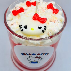 Puede incluir: Una vela roja de Hello Kitty con una cobertura de crema batida blanca y una figurita de Hello Kitty en la parte superior. La vela tiene una etiqueta blanca con las palabras "Hello Kitty" y una imagen de dibujos animados de Hello Kitty.