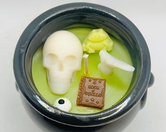 May include: A black cauldron-shaped candle with a light green wax interior. The candle features a white skull, a yellow frog, a white bone, a brown book with the text "Book of Spells", and a white eyeball. The candle is designed for Halloween.