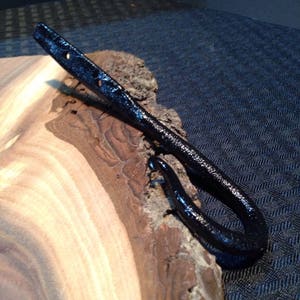 May include: Black metal hook with a rounded loop, mounted on a wooden slab with a natural wood grain pattern.