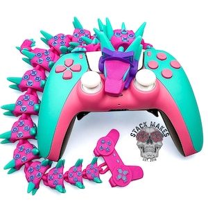 May include: A custom-designed video game controller in pink and teal, adorned with a dragon-like embellishment. Additional decorative pieces are scattered around the controller. The controller has white joysticks and pink buttons. The logo "STACK MAKES" is visible.