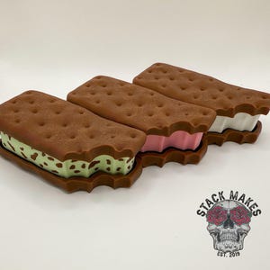May include: Three ice cream sandwich-shaped containers with brown 'cookie' tops and different colored 'ice cream' fillings: green with brown spots, pink, and white. The containers are on a white background. The logo "STACK MAKES EST. 2019" is in the bottom right corner.
