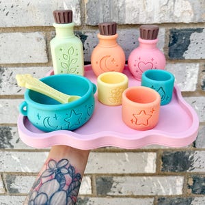 May include: A set of silicone play items on a light pink cloud-shaped tray, including a teal cauldron with a yellow spoon, three bottles with brown caps, and three small cups in yellow, orange, and teal, all featuring embossed celestial and floral motifs.