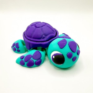 May include: A 3D-printed turtle toy in bright colors. The turtle has a purple shell and purple spots on its turquoise head and feet. The turtle's eye is large and white with a black pupil. The toy is designed with a textured surface.