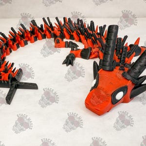May include: A 3D printed red and black dragon with a spiky back and a white eye. The dragon is posed with its head tilted to the side and its mouth slightly open.