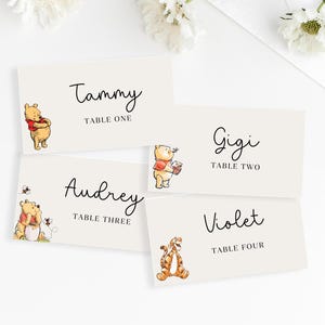 May include: Four cream-colored place cards with Winnie the Pooh characters. Each card has a name in black script and the table number. Winnie the Pooh, Piglet, and Tigger are featured on the cards. White flowers are in the background.