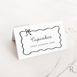 May include: White card with a black border and a handwritten sign that says "Cupcakes" with the flavors "Cheese, Chocolate, Lemon" listed below.