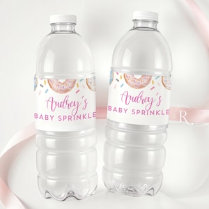 May include: Two clear plastic water bottles with white caps. Each bottle has a label with a pink donut design and the text "Audrey's Baby Sprinkle". A pink ribbon is in the background. The bottles are for a baby shower.