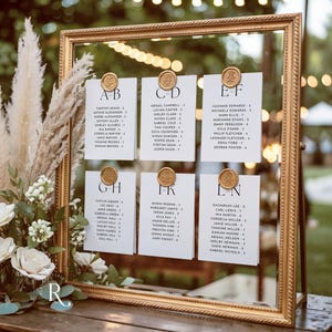 May include: A gold framed seating chart with white cards, each labeled with a letter combination and names. The cards are affixed with gold wax seals. The background includes flowers and greenery, suggesting a wedding or event.