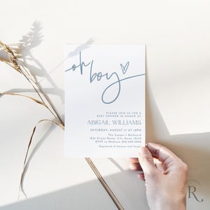 May include: A white baby shower invitation with blue text. The words "oh boy" are written in a cursive font, with a heart symbol. The invitation includes details for Abigail Williams' baby shower, including the date, time, and location.