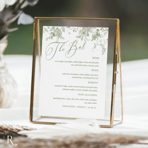 May include: A gold-framed sign with a printed menu for a bar, featuring categories for beer, wine, spirits, and non-alcoholic beverages. The menu includes a floral design and the text "The Bar".