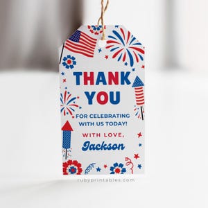 May include: White gift tag with red, white, and blue patriotic graphics, including an American flag, fireworks, and floral designs. The tag reads "THANK YOU" and "FOR CELEBRATING WITH US TODAY!" with the name "Jackson" at the bottom.