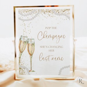 Könnte beinhalten: Ein weißes Schild mit goldener Schrift, auf dem "Pop the Champagne She's Changing Her Last Name" steht. Das Schild ist mit goldenem Glitzer und weißen Perlen verziert. Zwei Champagnergläser sind auf dem Schild abgebildet.
