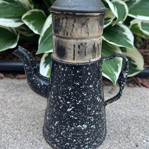 Può includere: Una cafetera vintage in smalto nero e bianco macchiettato con un coperchio rimovibile e un filtro in metallo. La caffettiera ha un aspetto usurato e invecchiato.