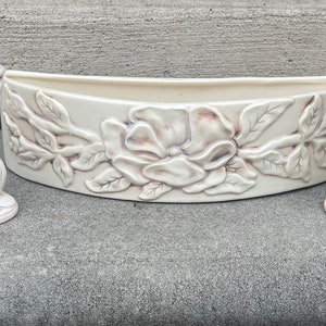 Puede incluir: Un juego de tres candelabros de cerámica blanca con un diseño floral. La pieza central es una maceta en forma de media luna con una flor de magnolia y hojas en relieve.