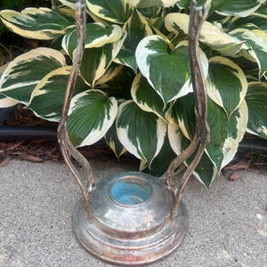 May include: A silver-toned metal candle holder with two curved, branch-like handles. The base is round with a blue interior. The candle holder is placed on a gray concrete surface, with green and white leafy plants in the background.