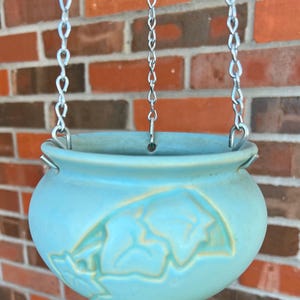 Op de afbeelding: Een hangende turquoise keramische plantenbak met een bladeren ontwerp, opgehangen aan zilveren kettingen. De ronde pot heeft een gladde afwerking en staat tegen een bakstenen muur. Een decoratief stuk voor planten.