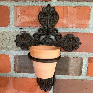 May include: A black metal wall planter shaped like a cross with a terracotta pot. The planter is mounted on a brick wall.
